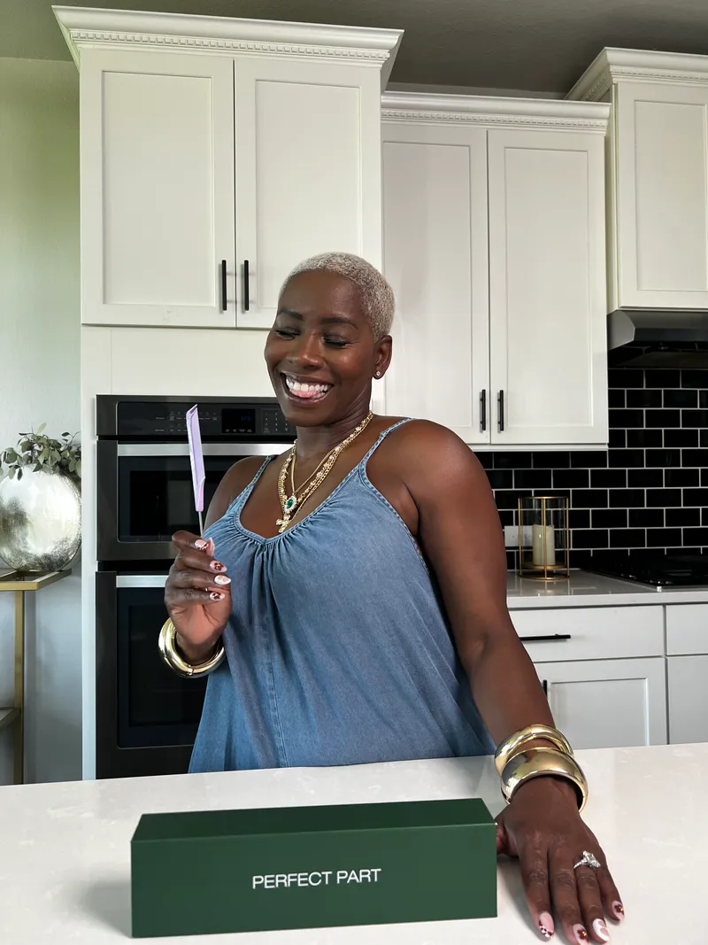 Monique smiling in her kitchen holding a PERFECT PART comb with the signed green box on the counter